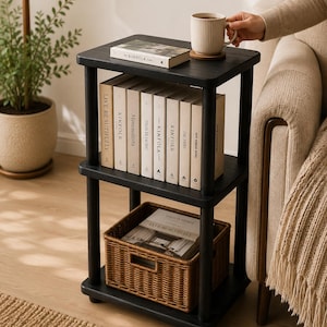 May include: A black three-tier side table with four cylindrical legs. The top shelf holds a book and a ribbed mug. The middle shelf contains a row of books with white spines. The bottom shelf features a rectangular brown woven wicker basket.