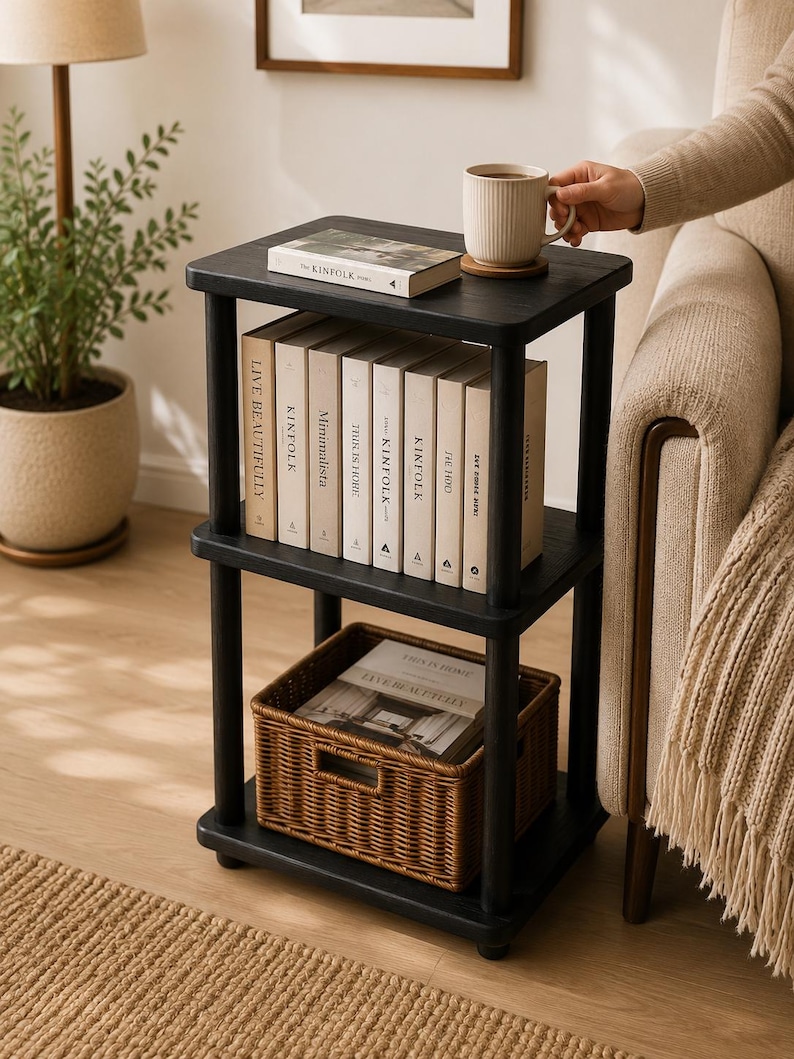 May include: A black three-tier side table with four cylindrical legs. The top shelf holds a book and a ribbed mug. The middle shelf contains a row of books with titles including Kinfolk and Minimalista. The bottom shelf holds a woven basket.