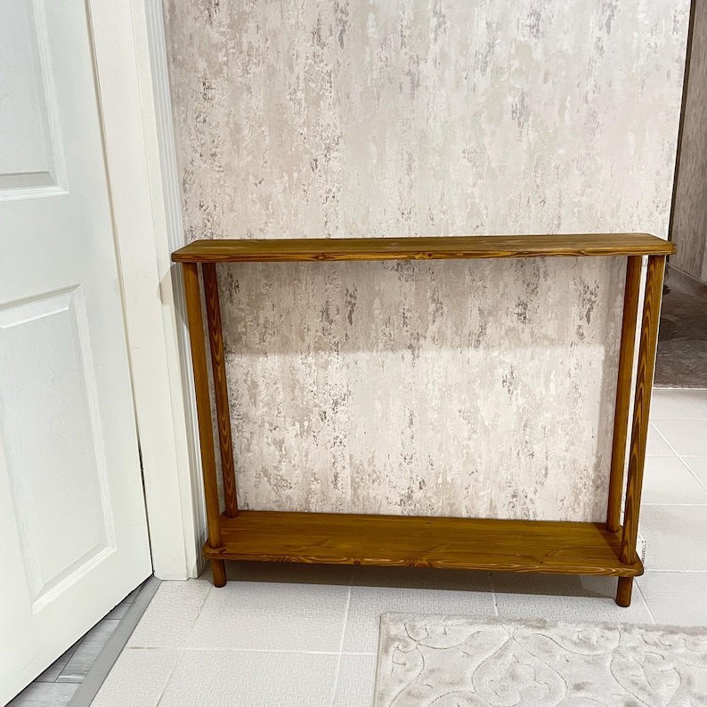 Op de afbeelding: Een houten consoletafel met twee planken. De tafel is gemaakt van natuurlijk hout en heeft een rustieke afwerking. De tafel staat voor een witte muur.