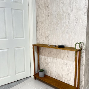 Op de afbeelding: Een houten consoletafel met een plank, met een natuurlijke houten afwerking. De tafel staat in een kamer met witte muren en een witte deur. Er staan twee kleine potplanten op de tafel.
