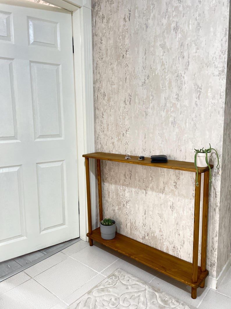 Op de afbeelding: Een houten consoletafel met een plank, met een natuurlijke houten afwerking. De tafel staat in een kamer met witte muren en een witte deur. Er staan twee kleine potplanten op de tafel.