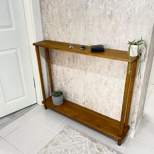 Op de afbeelding: Een houten consoletafel met twee planken. De bovenste plank is leeg, terwijl de onderste plank een kleine plant in een pot heeft. De tafel is gemaakt van donkerbruin hout en heeft een eenvoudig, modern ontwerp.