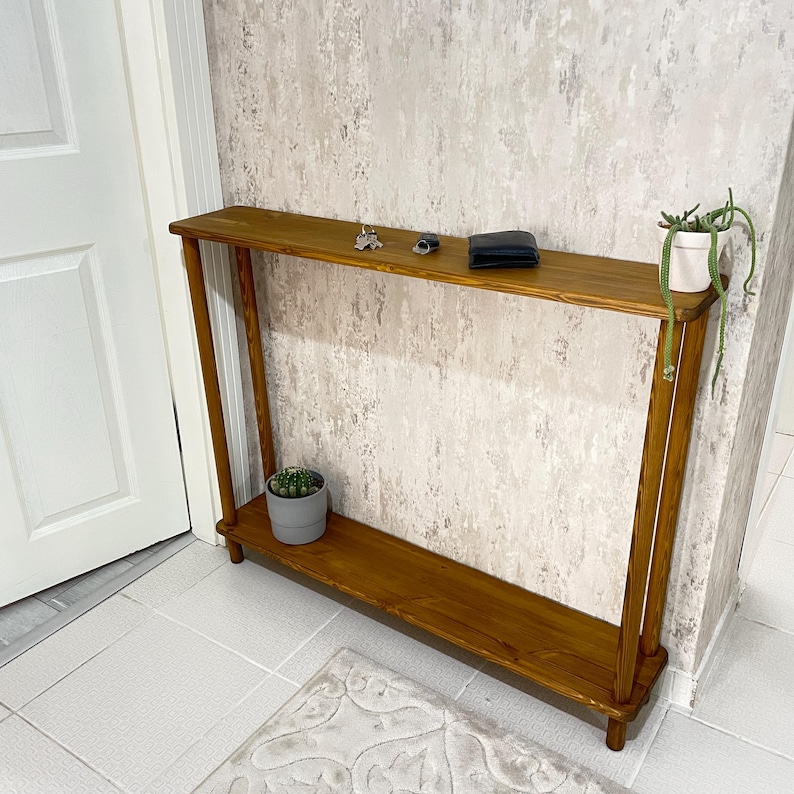 Op de afbeelding: Een houten consoletafel met twee planken. De bovenste plank is leeg, terwijl de onderste plank een kleine plant in een pot heeft. De tafel is gemaakt van donkerbruin hout en heeft een eenvoudig, modern ontwerp.