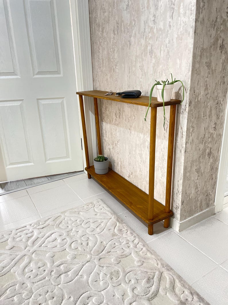 Op de afbeelding: Een houten consoletafel met twee planken. De tafel is in een lichte bruine kleur gebeitst en heeft een eenvoudig ontwerp. Een kleine plant in een pot staat op de bovenste plank.