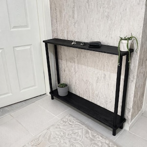 May include: A black wooden console table with two shelves. The table has a rectangular top and a lower shelf, supported by black legs. Small potted plants and a wallet are on the top shelf. The table is placed against a textured wall.