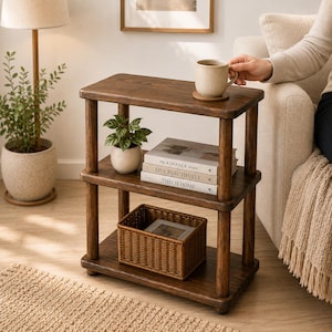 Op de afbeelding: Een houten bijzettafel met drie niveaus en een donkerbruine afwerking. Op de bovenste plank staat een geribbelde keramische mok. De middelste plank bevat een kleine potplant en drie gestapelde boeken met de titels The Kinfolk Home, Live Beautifully en This Is Home.