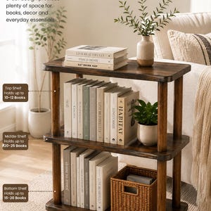 May include: A three-tier wooden bookshelf with a dark brown finish. The top shelf holds three books and a vase with greenery. The middle shelf contains several books and a small potted plant. The bottom shelf holds books and a woven basket.