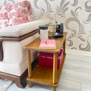 Op de afbeelding: Een houten bijzettafel met twee planken. De bovenste plank heeft een notitieboek, een pen en een zwarte portemonnee. De onderste plank heeft een roze doos. De tafel staat voor een witte bank met een kussen met bloemenpatroon.