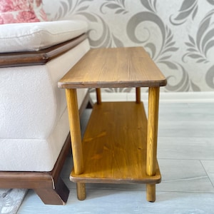 Könnte beinhalten: Ein kleiner Holz-Beistelltisch mit zwei Ablagen. Der Tisch ist in einem hellen Braunton gebeizt und hat ein schlichtes Design.