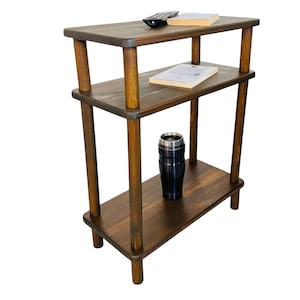 May include: A three-tiered, dark brown wooden side table with rounded corners and cylindrical legs. A black travel mug, a remote control, and two books are on the shelves. The table is designed for storage and display.