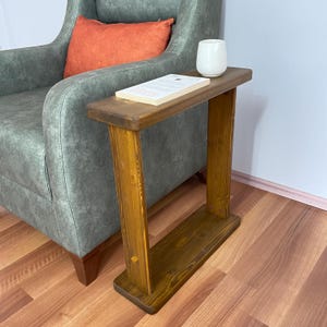 May include: A small, wooden side table with a rectangular top and two vertical supports. The table is stained a warm brown color. A white ceramic cup and a book are on the table. The table is next to a green armchair with an orange pillow.