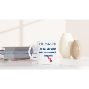 May include: White ceramic mug with a handle, featuring the text "WHAT IS GRAVITY?" and the humorous definition: "THE 'FALL GUY' WHO GETS BLAMED FOR EVERYTHING THAT GOES DOWN," with a red arrow graphic. The mug is on a white surface.