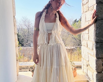 May include: A woman wearing a white, tiered maxi skirt and a matching halter top. The skirt has a ruffled hem and the top has a tie closure at the neck. The woman is standing in a doorway with a stone archway.