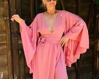 May include: A flowing, light pink dress with wide sleeves and a wrap-around waist. The dress features a deep V-neckline and a high slit on the side. The model is wearing sunglasses and sandals. The background includes a wooden door and greenery.