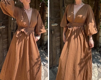 May include: A long, flowing brown dress with a deep V-neckline and wide sleeves. The dress features a matching fabric belt that cinches the waist. The dress is made of a textured fabric and has a relaxed, bohemian style.