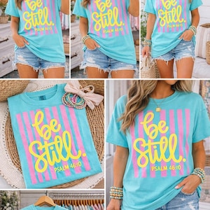 May include: A light blue t-shirt with the words "Be Still" in yellow script, set against pink vertical stripes. The text "PSALM 46:10" is below. The shirt is paired with denim shorts.