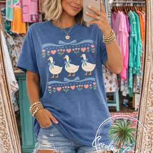 May include: A blue t-shirt featuring three white geese with blue bows, set against a decorative border of pink hearts, flowers, and blue bows. The shirt has a vintage wash appearance.