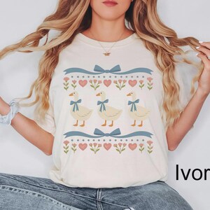 May include: Ivory-colored t-shirt featuring a whimsical design. The design includes three cartoon geese with blue bow ties, surrounded by blue ribbons, hearts, and floral elements. The word "Ivory" is printed on the bottom right.