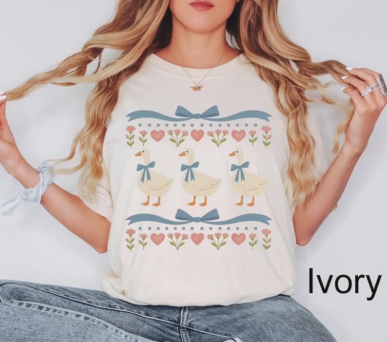 May include: Ivory-colored t-shirt featuring a whimsical design. The design includes three cartoon geese with blue bow ties, surrounded by blue ribbons, hearts, and floral elements. The word "Ivory" is printed on the bottom right.