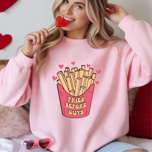 Op de afbeelding: Een lichtroze sweatshirt met een afbeelding van een frietbakje en de tekst "Fries Before Guys" in een retro lettertype. Het ontwerp bevat kleine roze hartjes. De sweatshirt is een casual, comfortabele stijl.
