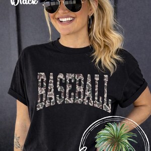 May include: Black t-shirt with the word "BASEBALL" in a camouflage pattern. The model is wearing a white baseball cap with "BASEBALL MOM" printed on it and sunglasses. The words "Comfort Colors Black" are in the upper left corner.