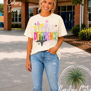May include: An ivory short-sleeve t-shirt featuring a graphic print with the text "Make A Difference" in colorful, stylized lettering. The design includes yellow, black, and pink star shapes along with multi-colored paint splatter accents.