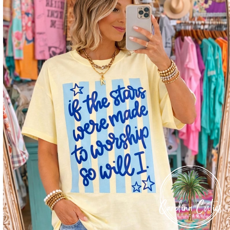May include: A light yellow t-shirt with the text "if the stars were made to worship so will I." in blue. The text is on a light blue striped background. The shirt has short sleeves and a crew neck.