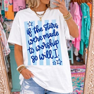 May include: White t-shirt with blue vertical stripes and the text "if the stars were made to worship so will I." The shirt has a relaxed fit and short sleeves. The model is wearing denim shorts.