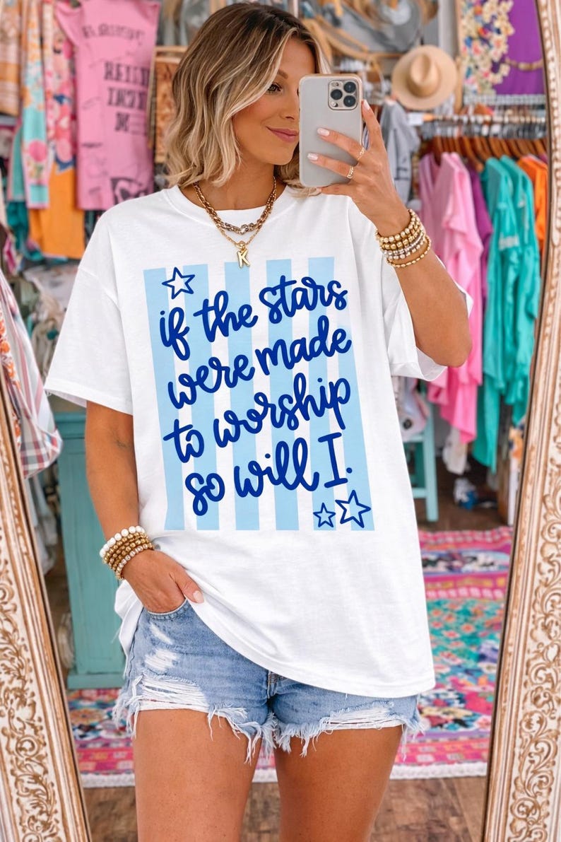 May include: White t-shirt with blue vertical stripes and the text "if the stars were made to worship so will I." The shirt has a relaxed fit and short sleeves. The model is wearing denim shorts.