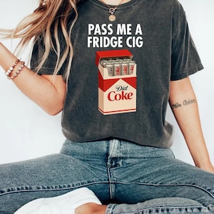 May include: Dark gray t-shirt with the text "PASS ME A FRIDGE CIG" in white. The shirt features a graphic of a Diet Coke carton filled with Diet Coke cans. The person is wearing blue jeans and white sneakers.