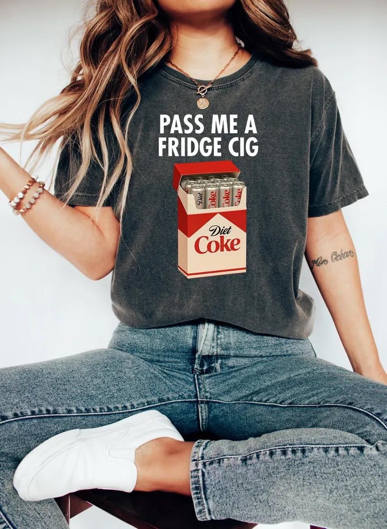 May include: Dark gray t-shirt with the text "PASS ME A FRIDGE CIG" in white. The shirt features a graphic of a Diet Coke carton filled with Diet Coke cans. The person is wearing blue jeans and white sneakers.