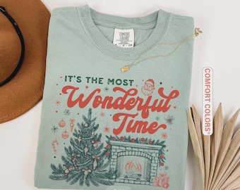 May include: A light green t-shirt with the phrase "It's the Most Wonderful Time" in red script. The graphic includes a Christmas tree, fireplace, and Santa Claus. A gold necklace is draped across the shirt.