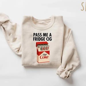 May include: Sand-colored sweatshirt with the text "PASS ME A FRIDGE CIG" above a graphic of a Diet Coke cigarette pack. The pack is red and white, with silver Diet Coke cans inside. The sweatshirt has long sleeves.