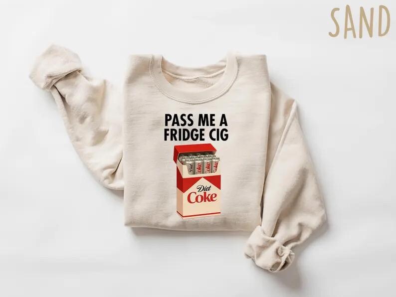 May include: Sand-colored sweatshirt with the text "PASS ME A FRIDGE CIG" above a graphic of a Diet Coke cigarette pack. The pack is red and white, with silver Diet Coke cans inside. The sweatshirt has long sleeves.
