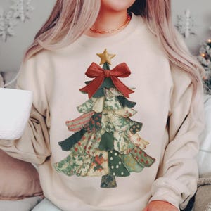 May include: Cream-colored sweatshirt featuring a patchwork Christmas tree design. The tree is adorned with a red bow, a gold star, and various fabric patterns in green, red, and cream. A festive and cozy apparel item.