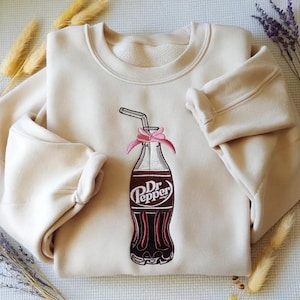 Peut inclure: Sweat-shirt crème avec un motif brodé de bouteille de Dr. Pepper. La bouteille est marron avec le logo Dr. Pepper et un nœud rose. Le sweat-shirt a un col rond et des manches longues.