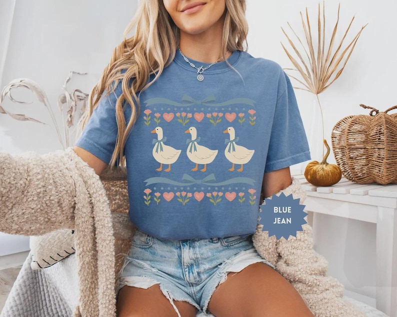 May include: Blue jean t-shirt featuring a whimsical design of three geese with blue ribbons around their necks. The geese are set against a backdrop of hearts, flowers, and ribbons. The shirt is a comfortable, casual style.