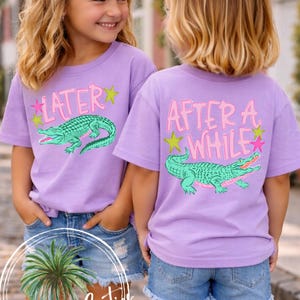 May include: Two young girls wearing matching lavender t-shirts with alligator graphics and text. One shirt says "LATER" and the other says "AFTER A WHILE". Both girls wear denim shorts and white bows. The shirts have pink and yellow accents.