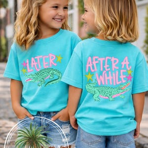May include: Two young girls wearing matching turquoise t-shirts with alligator graphics and the words "LATER" and "AFTER A WHILE" in pink. They are also wearing denim shorts and white bows in their hair. The shirts are from Carolina Cuties Boutique.