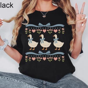May include: Black crew neck t-shirt with a whimsical design. The design features three geese with blue bows, surrounded by hearts, flowers, and bows. The word "Black" is printed at the top left.