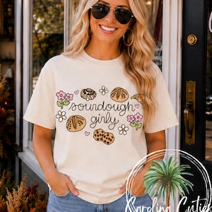 May include: A cream-colored short-sleeve t-shirt featuring a graphic print of various baked goods, including bread loaves, a cinnamon roll, a croissant, and cookies, interspersed with small pink hearts and colorful flowers, with the text sourdough girly.