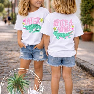 May include: Two young girls wearing white t-shirts with a pink and green alligator graphic and the text "LATER" and "AFTER A WHILE". They are wearing denim shorts and white sneakers, with white bows in their hair.