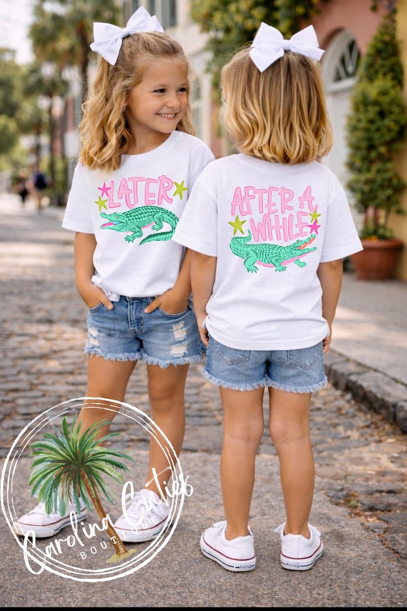 May include: Two young girls wearing white t-shirts with a pink and green alligator graphic and the text "LATER" and "AFTER A WHILE". They are wearing denim shorts and white sneakers, with white bows in their hair.