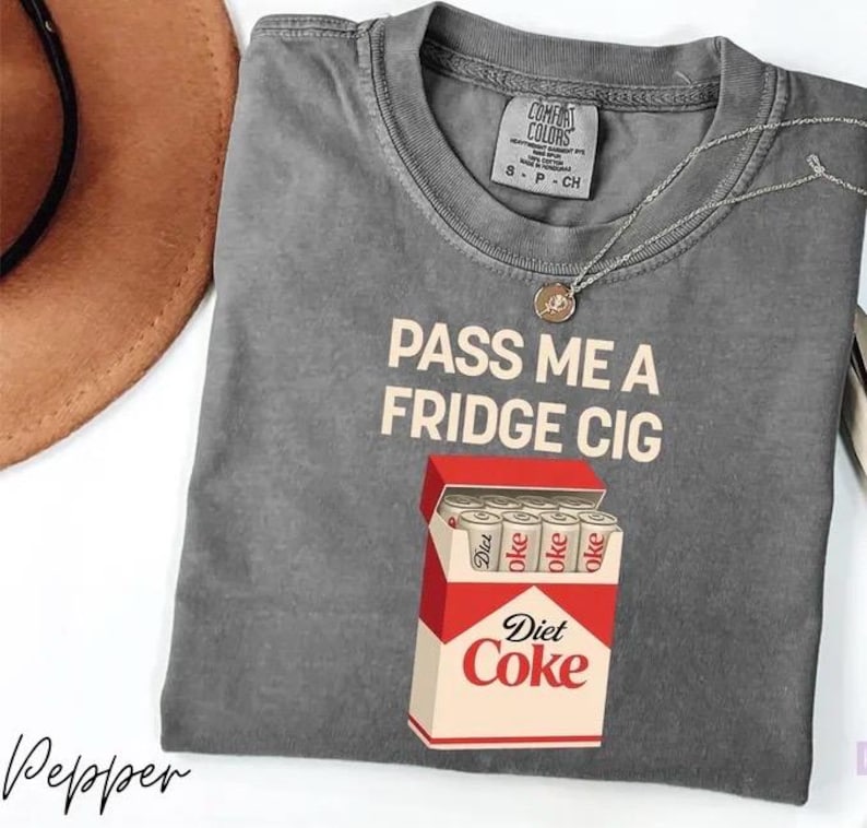 May include: Gray t-shirt with the text "PASS ME A FRIDGE CIG" and a graphic of a Diet Coke pack. The shirt has a crew neck and short sleeves. A silver necklace with a pendant is on the shirt.