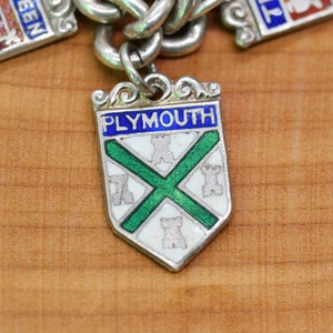 May include: Silver charm with enamel detailing in the shape of a shield. The shield is white with a green cross and four white castles. The word "PLYMOUTH" is written in blue enamel at the top of the shield.