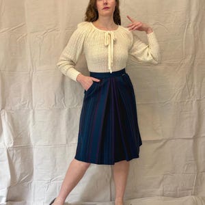May include: A woman wearing a white knitted sweater with a bow at the neckline and a dark blue and purple striped skirt.
