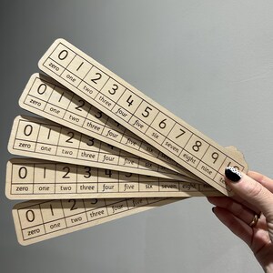 May include: Five light-coloured wooden number strips arranged in a fan shape. Each strip features a grid containing the digits zero through ten, with the corresponding English word written in lowercase letters directly below each numeral.