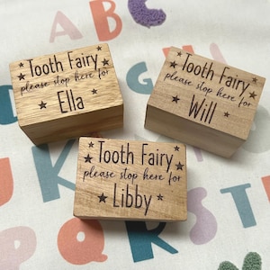 May include: Three wooden boxes with the text "Tooth Fairy please stop here for" followed by a name. The names are Ella, Will, and Libby. The boxes are on a colourful fabric background.