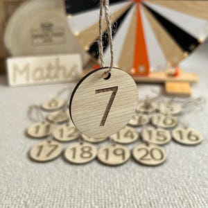 May include: A wooden number seven ornament with a jute string. The number is etched into the light-coloured wood. Other wooden number ornaments are visible in the background, along with a wooden wheel and the word "Maths".