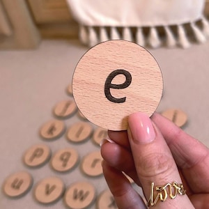 May include: A wooden disc featuring the lowercase letter "e" in black. Other wooden discs with letters are in the background. A gold ring with the word "love" is on a finger. A white fabric with the letters "VWXYZ" is in the background.
